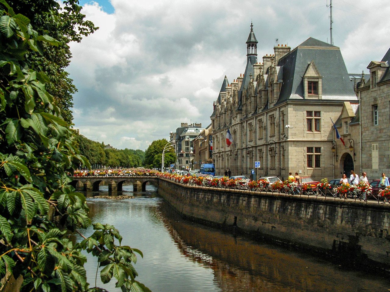 The Odet river in Quimper, Brittany: a stone bridge and a quay hung with red and white flowers, and the Finistère prefecture flying the French flag